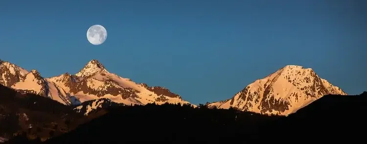 Snow-capped mountains under a clear sky with a visible full moon.
