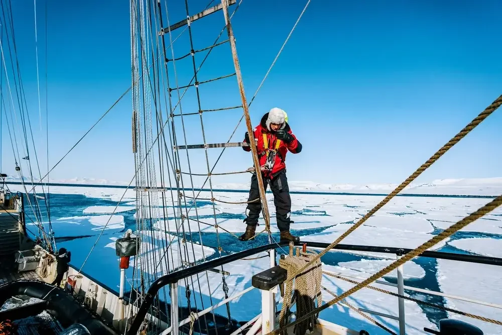 A person in winter clothing on a sailing ship navigating icy waters with floating ice and snow-covered landscape in the background.
