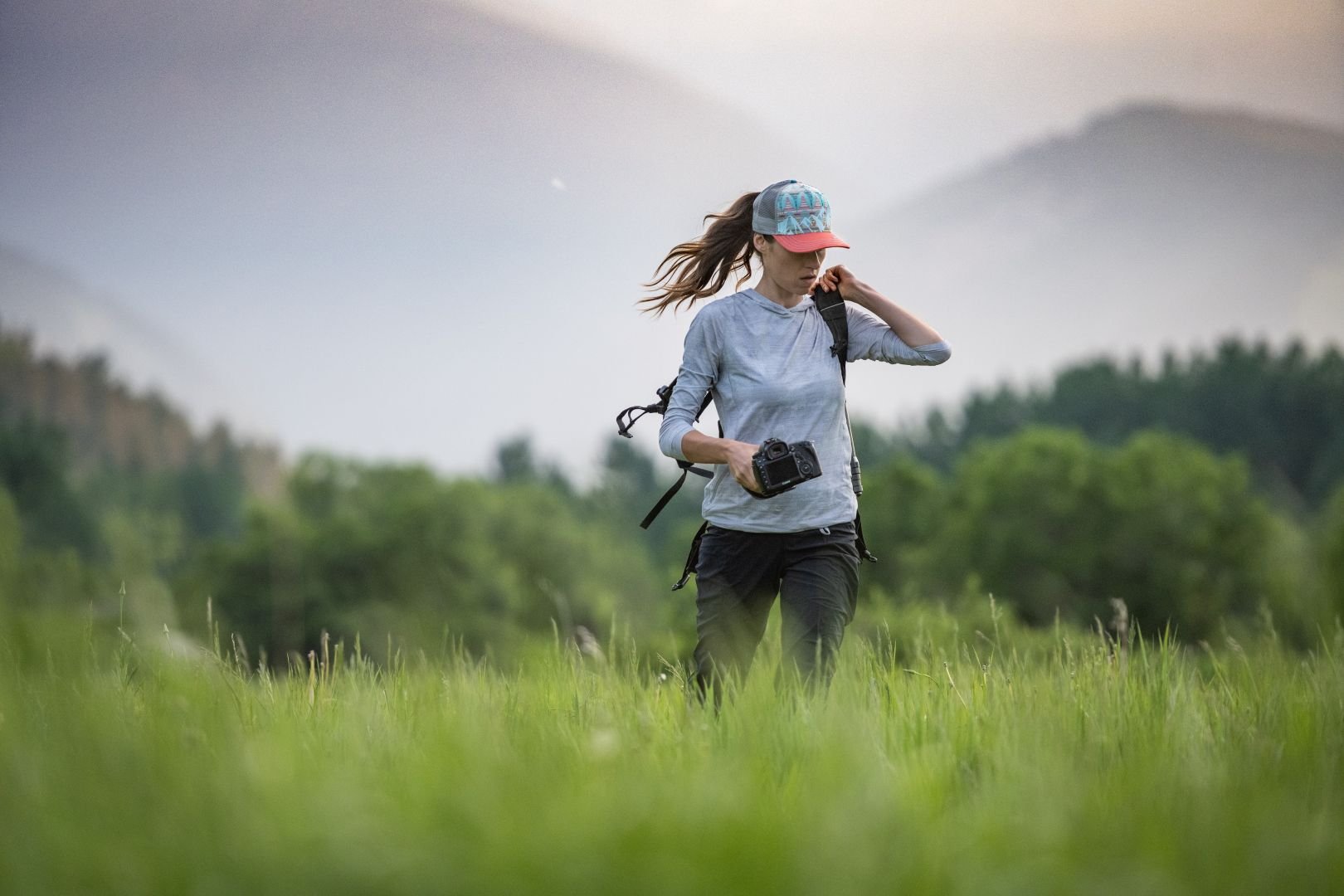 A woman walking through a grassy field with a camera in her hand, wearing a cap, long-sleeve shirt, and backpack, with trees and mountains in the background.