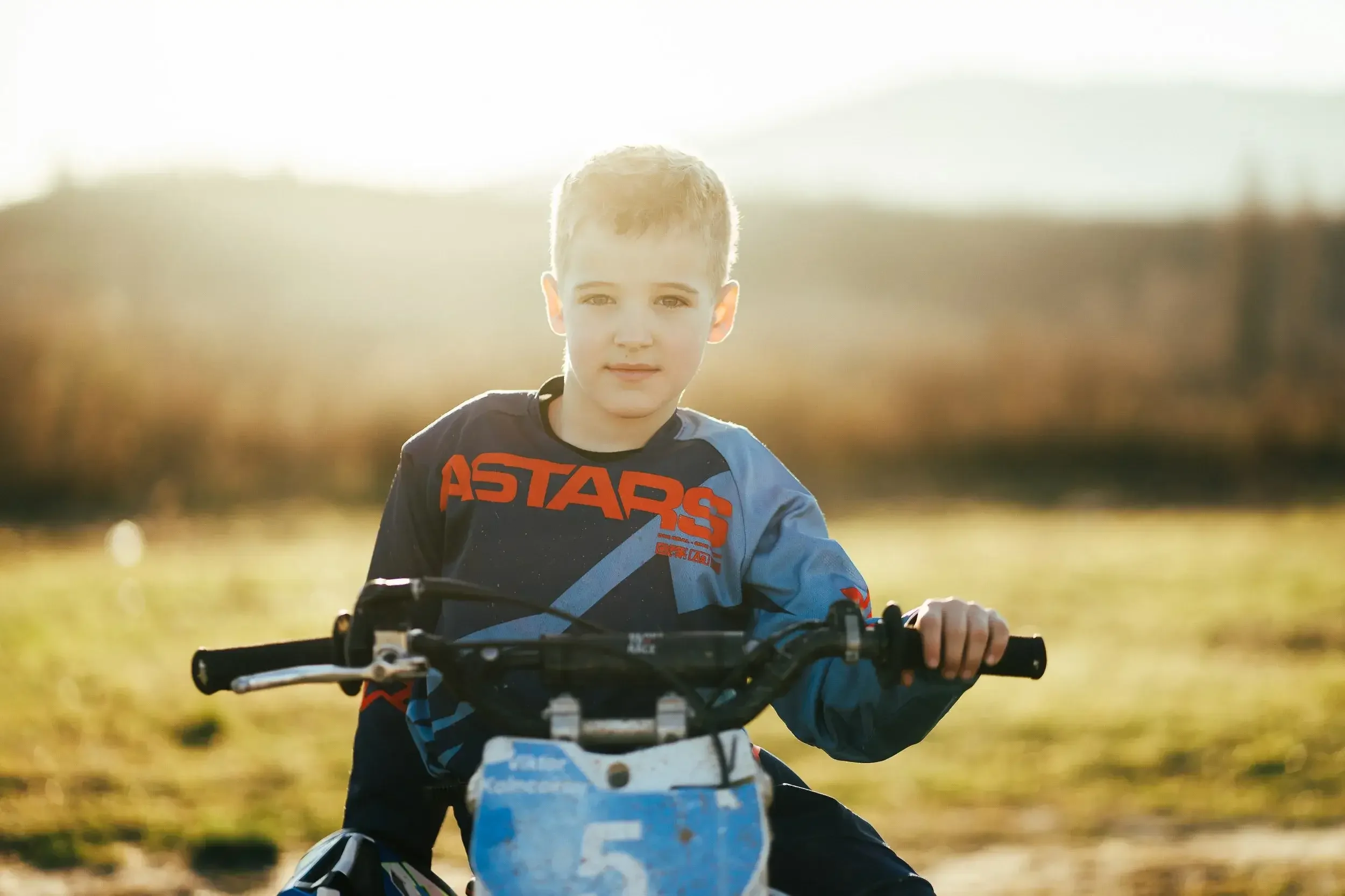 A young boy in a blue and red motocross jersey sits on a dirt bike outdoors during sunset, holding the handlebars and looking at the camera.
