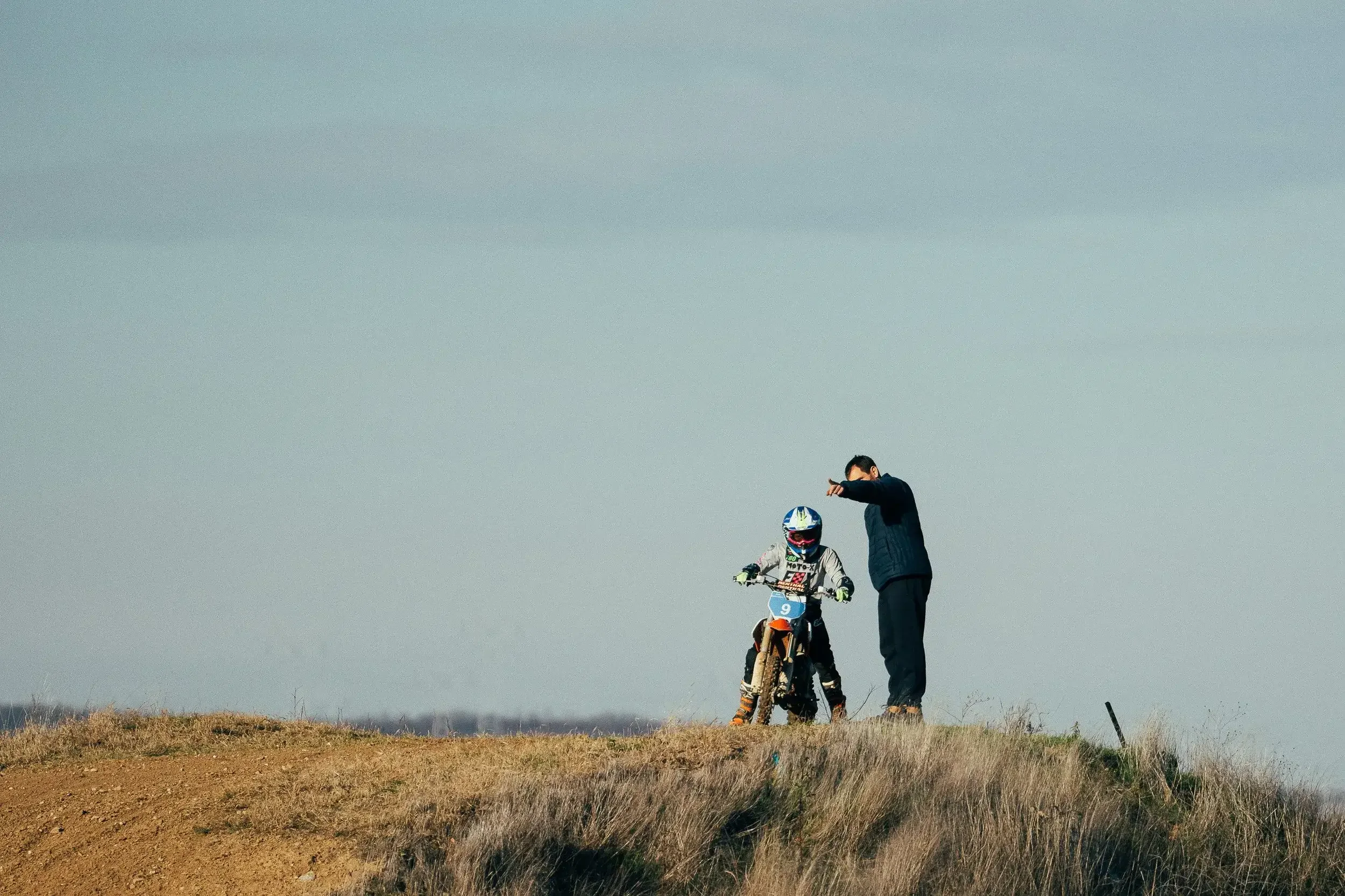 A young girl in motocross gear sitting on a dirt bike on a grassy hill, with an adult man standing next to her, pointing at the horizon, under a cloudy sky.
