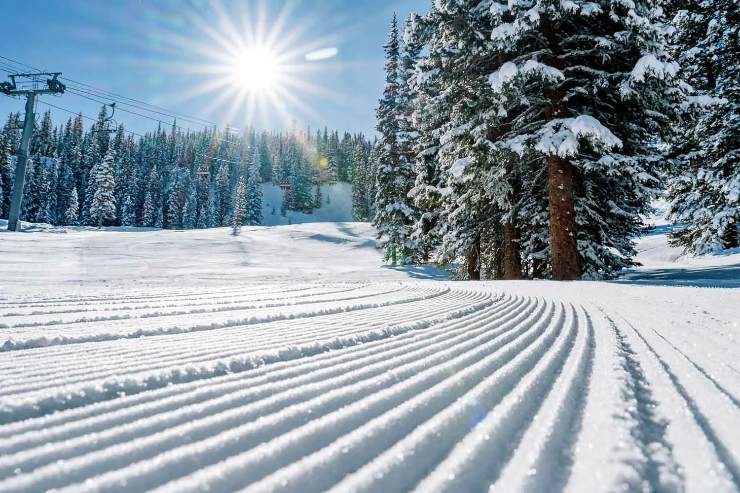 Snow-covered ski slope with groomed tracks, tall snow-laden evergreen trees, and a ski lift under a bright sun against a clear blue sky.