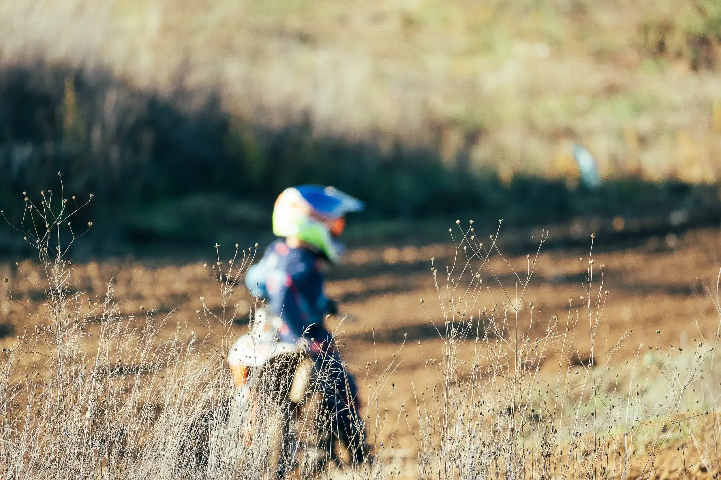 A child riding a dirt bike on a dirt path in outdoor natural surroundings, with dry grass and blurred greenery in the background.
