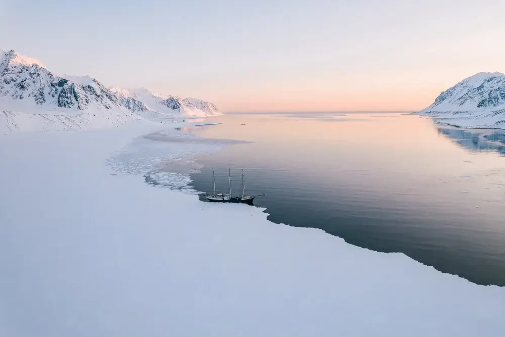 Aerial view of a glacier with a large iceberg in dark greenish water, and a small group of people on the snow-covered ice in the background.
