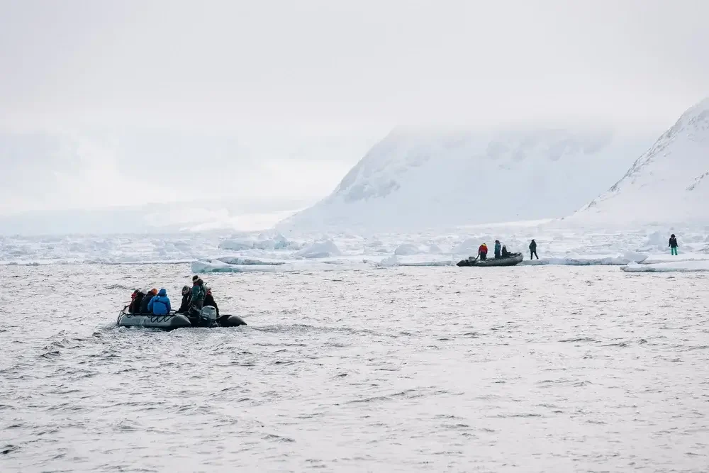 Group of people in life jackets on inflatable boats navigating icy Arctic waters with snow-covered mountains and icebergs in the background
