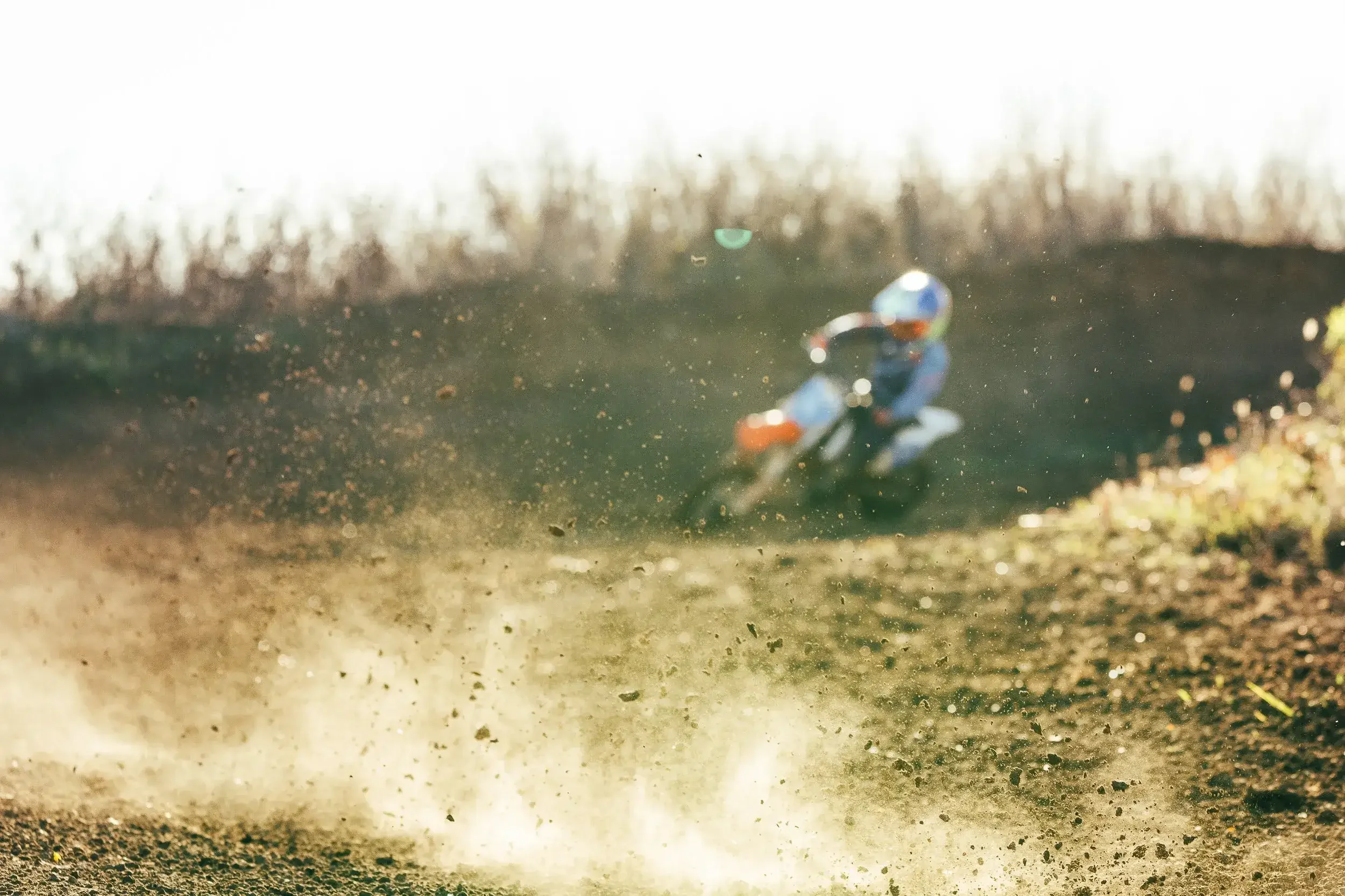 A motocross rider wearing a helmet and riding gear speeds around a dirt track, kicking up dirt and dust as they lean into a turn.

