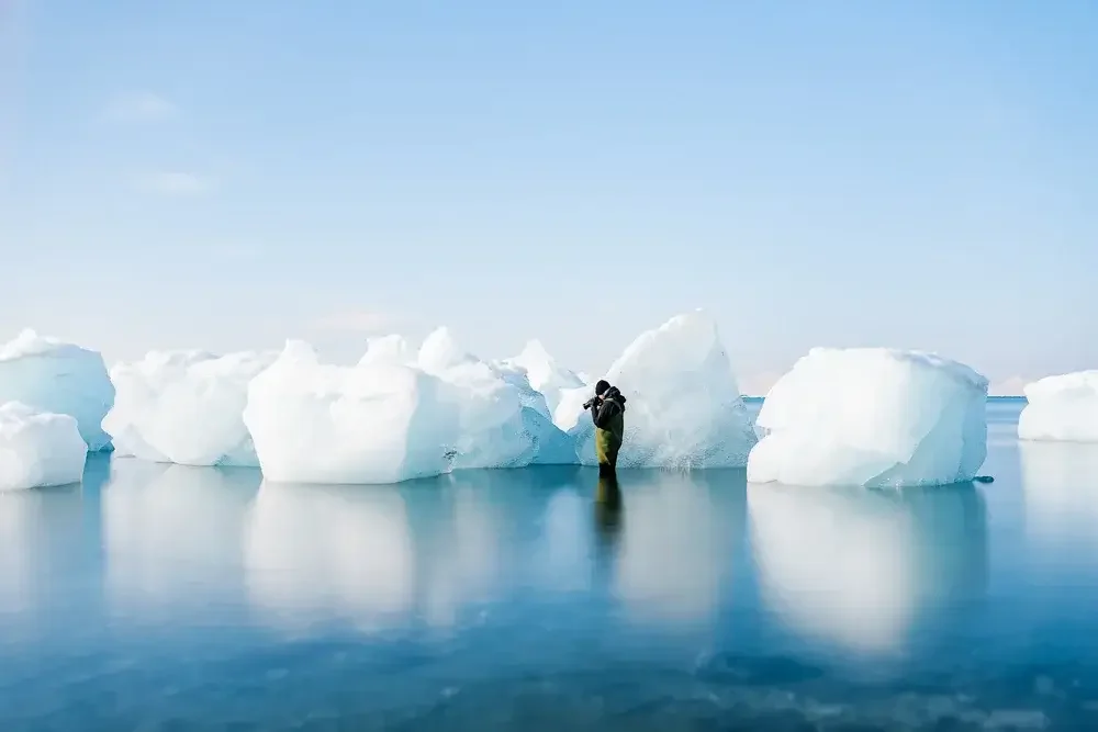 A person in a green and black jacket taking a photograph of icebergs floating in calm Arctic waters under a light blue sky.
