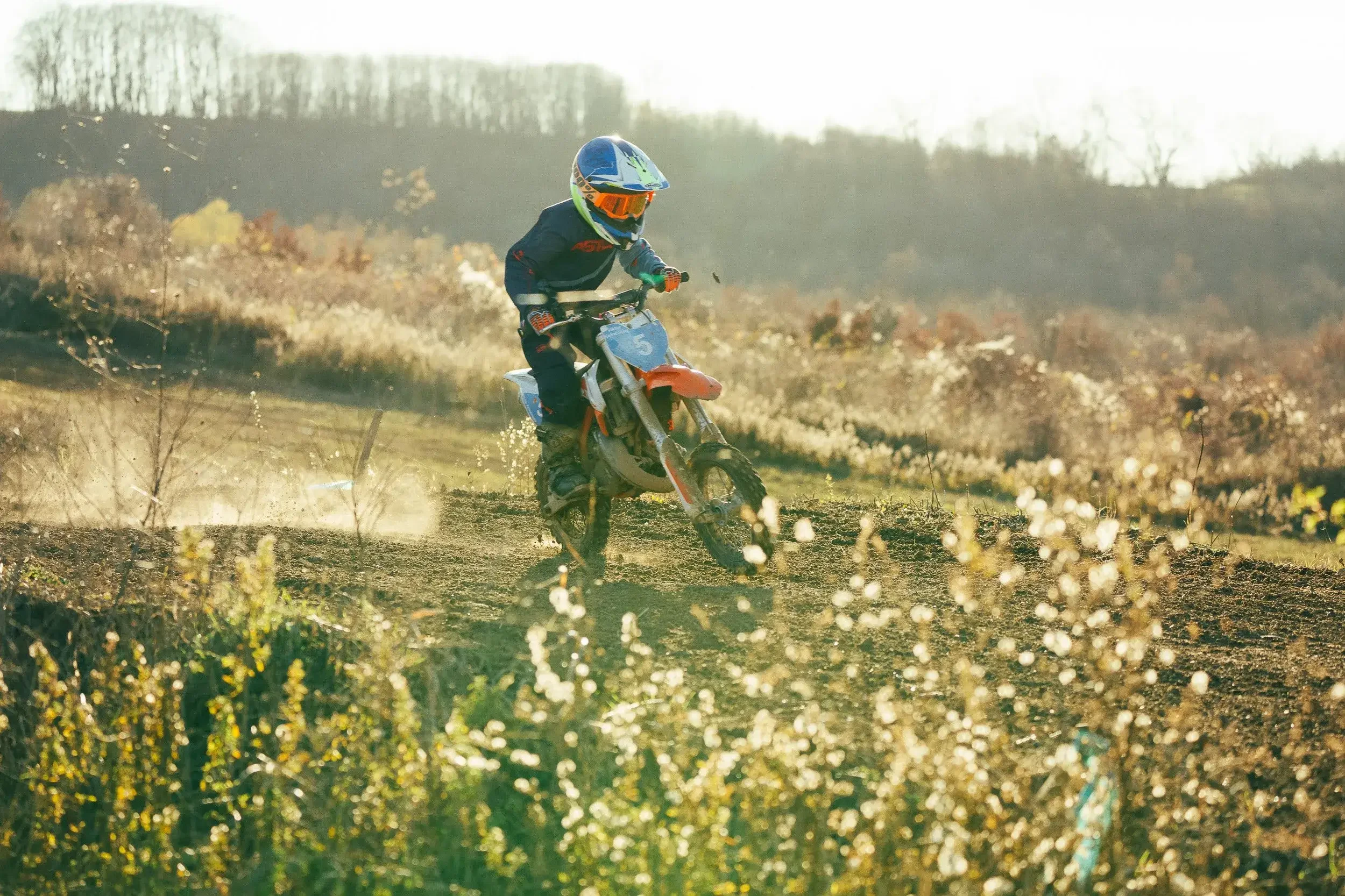A person wearing a helmet and riding a dirt bike on a trail in a rural area with dry vegetation and rolling hills in the background.
