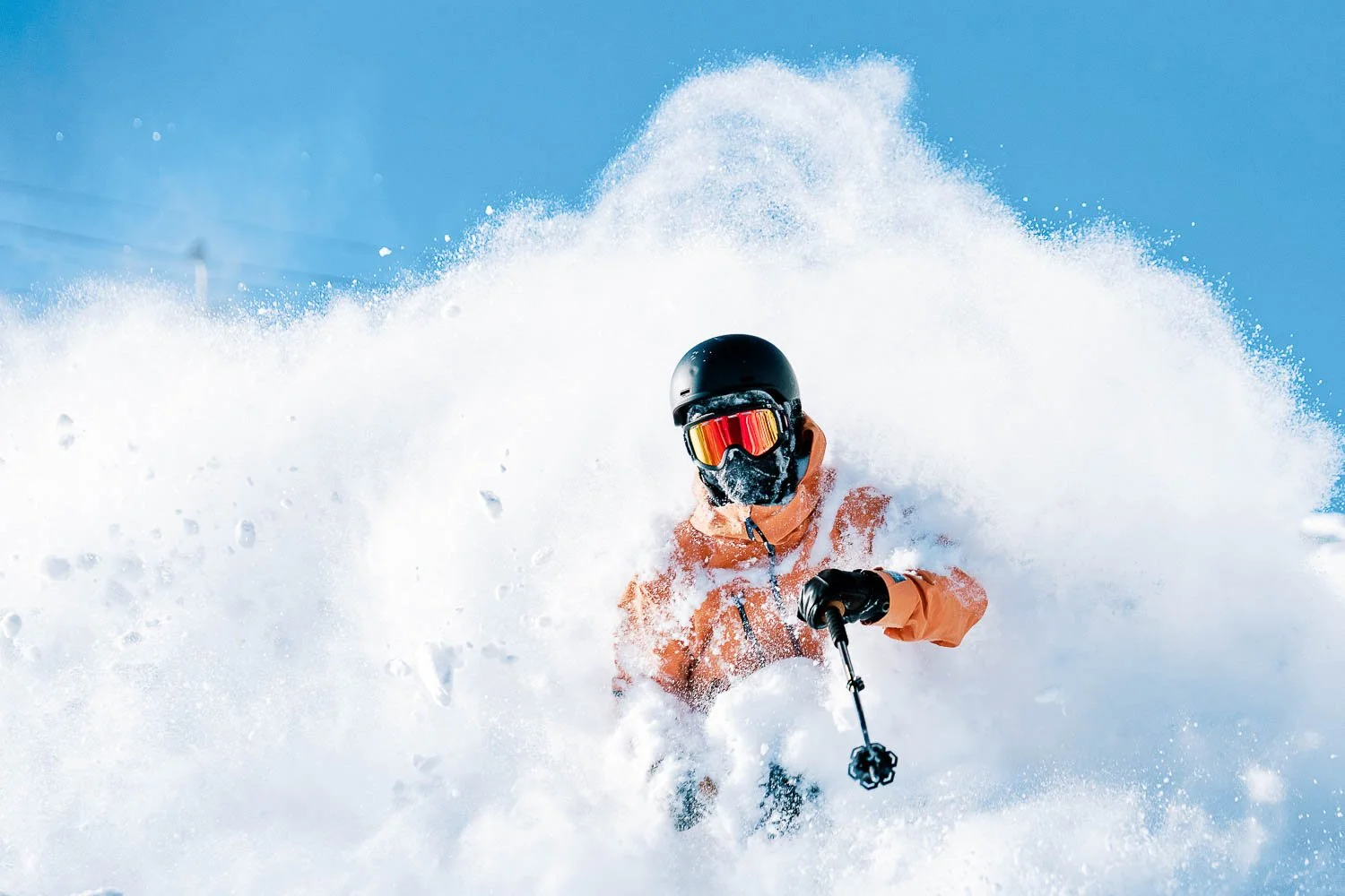 A person skiing in deep snow, wearing a black helmet, red goggles, and an orange jacket, with snow flying around.