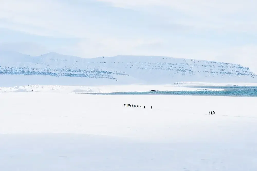 A sailboat in icy waters with snow-covered mountains in the background and a cloudy sky.
