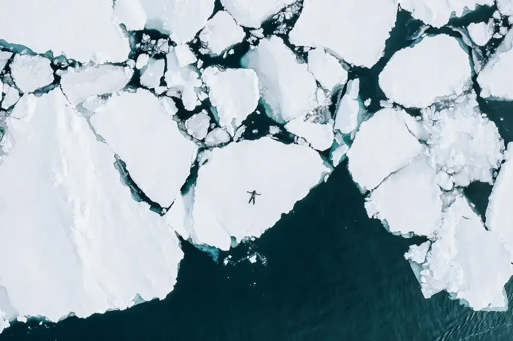 An aerial view of broken ice floes and a person lying on a large ice sheet surrounded by water in an Arctic or Antarctic setting.
