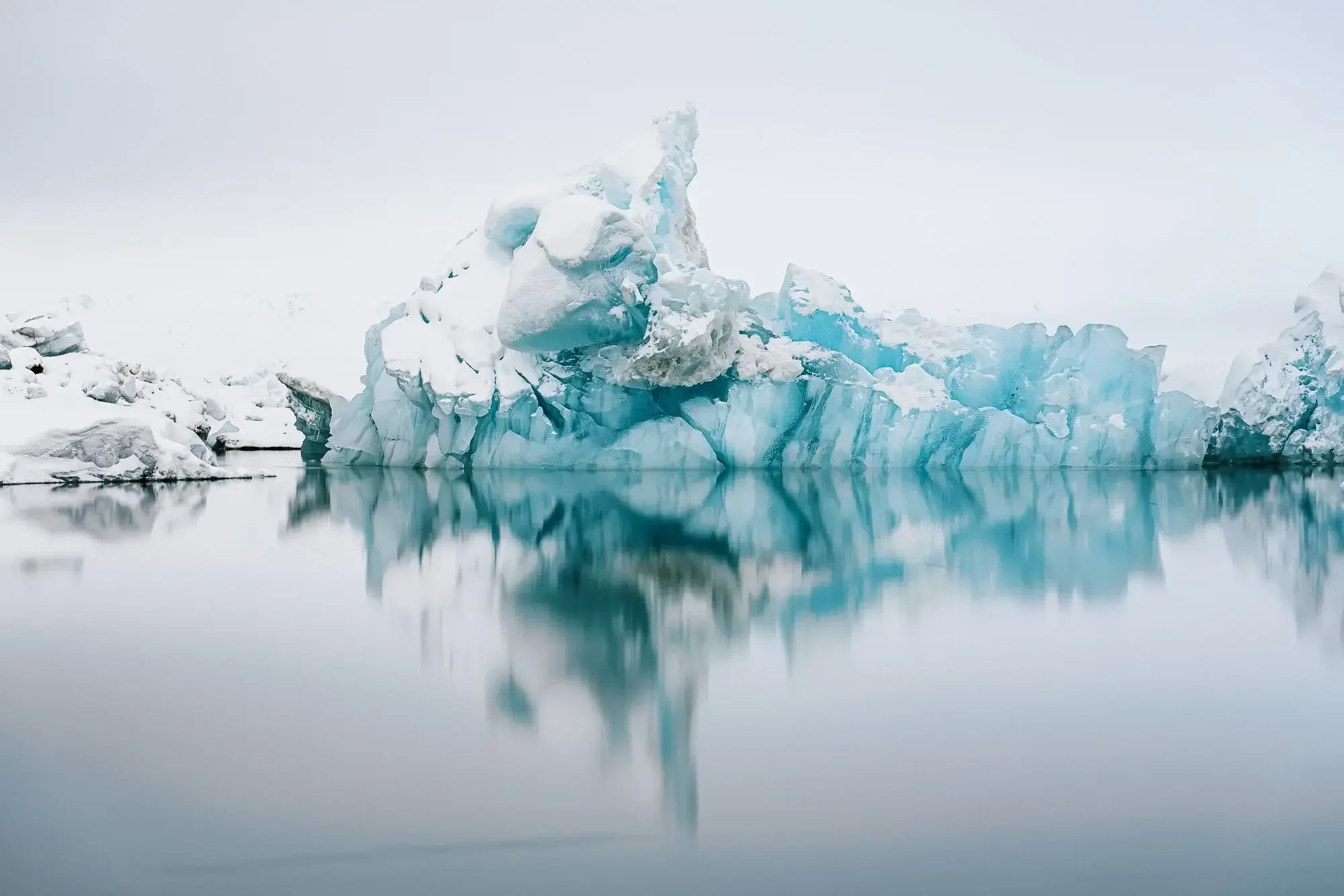 A large iceberg with snow-covered top floating in calm, reflective water in a cold, icy landscape.