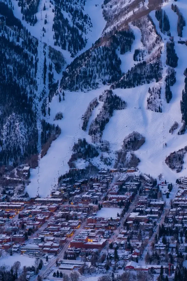 Aerial view of a snowy mountain town with ski slopes and forested mountains in the background.
