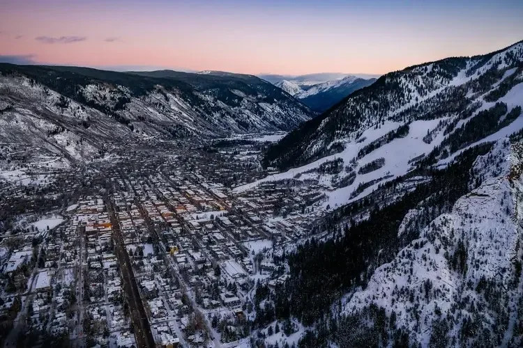 Snow-covered mountain ski resort with ski lifts and dense evergreen trees under a clear blue sky.

