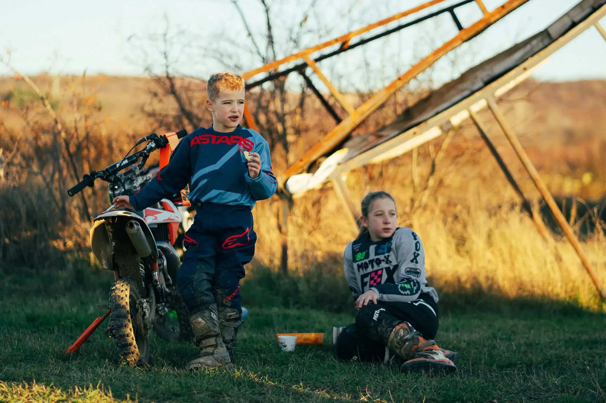 Two children, a boy and a girl, with motocross gear, taking a break outdoors near a fallen wind turbine with overgrown grass in the background.

