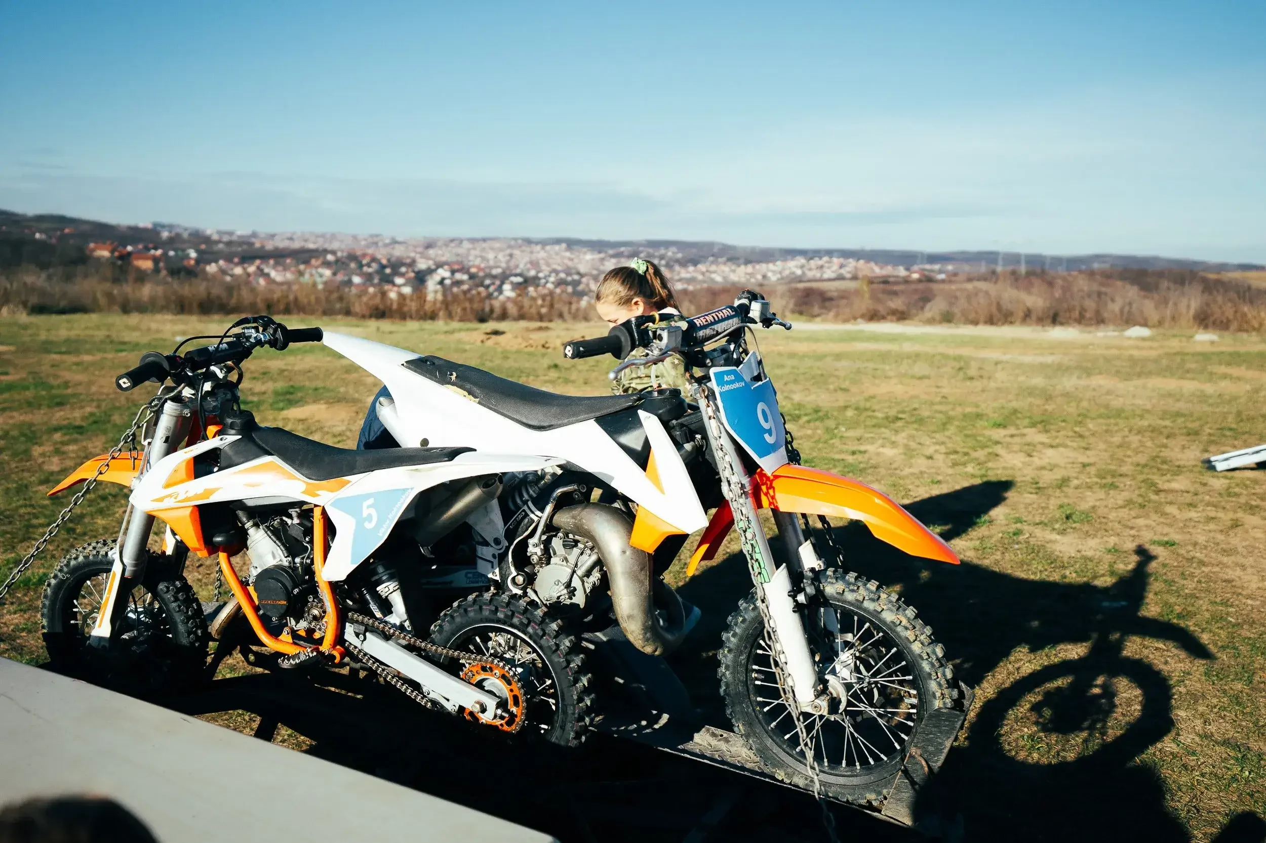 Two off-road motocross bikes positioned outdoors on a grassy area with a cityscape in the distance under a clear blue sky, one bike with a blue race number 9 plate and the other with a blue number 5 plate.
