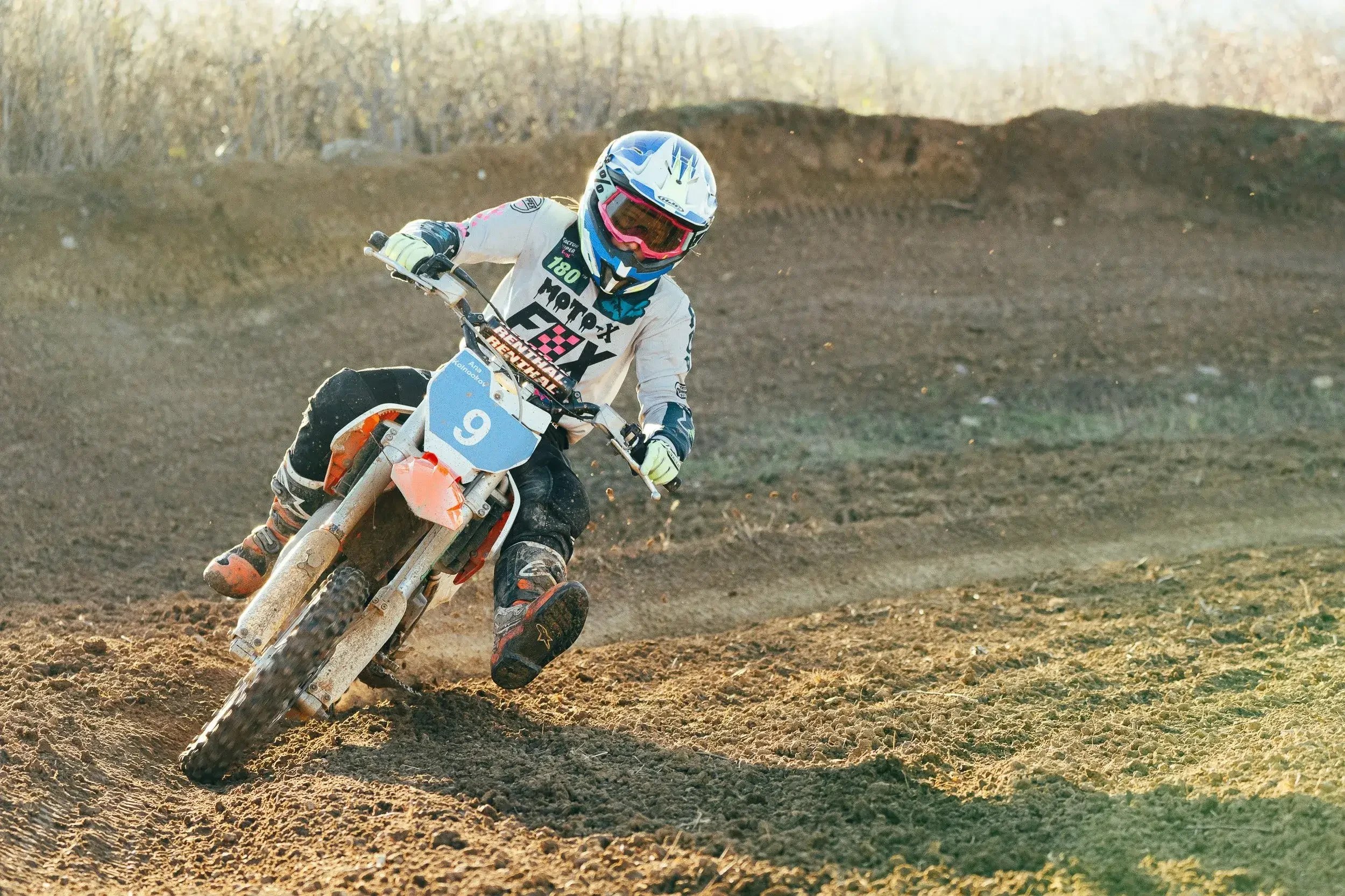 A motocross rider wearing a white and black suit and a blue and white helmet with goggles, riding a dirt bike on a rugged, dirt track with a background of leafless trees.
