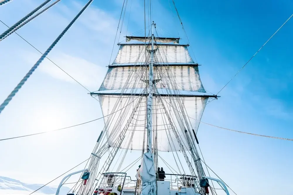 Looking up at the sails and rigging of a sailing ship against a blue sky.
