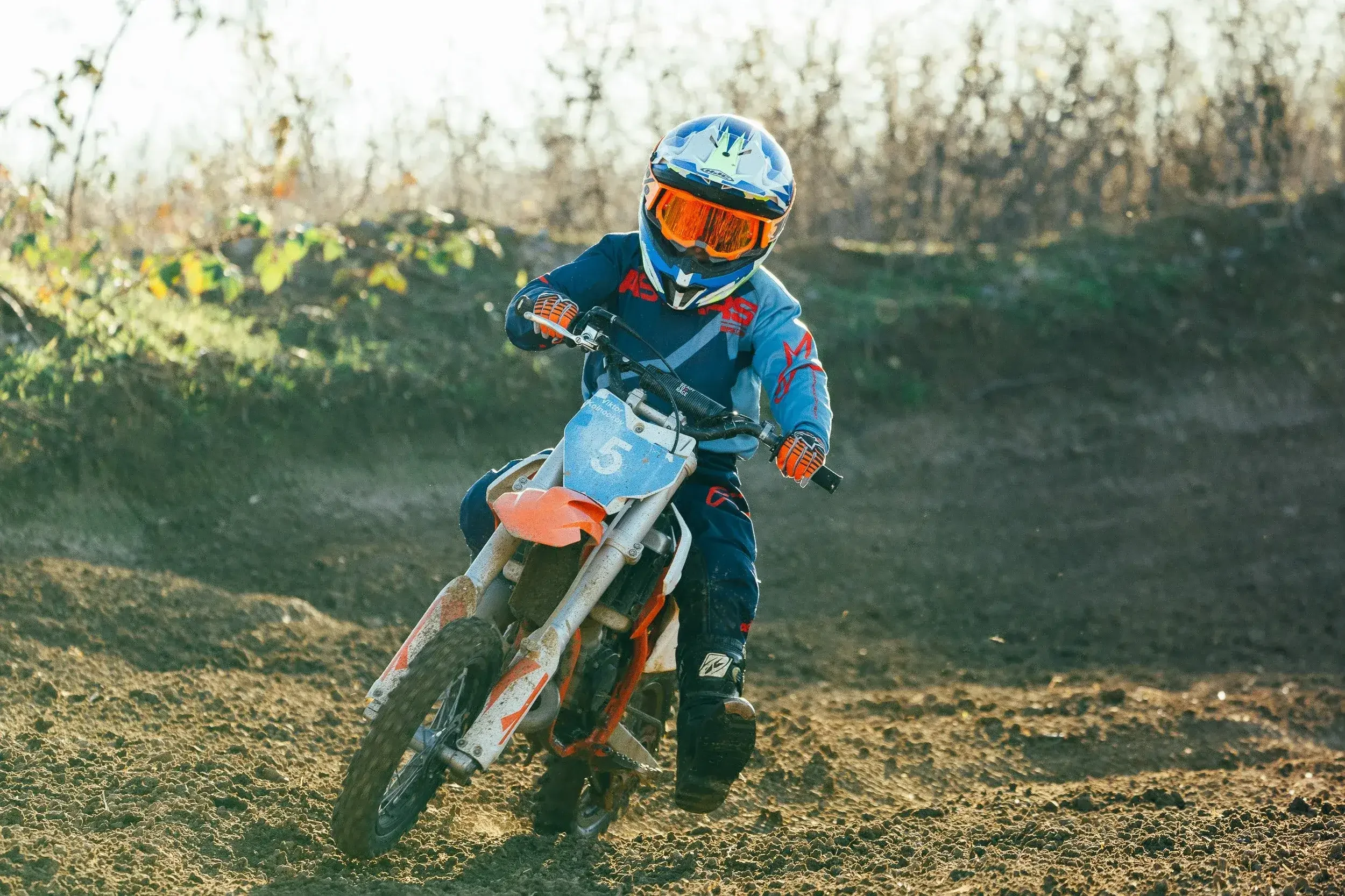 A young motocross rider wearing a blue and orange helmet and racing gear riding a dirt bike with the number 5 through a dirt track with a background of trees and cloudy sky.
