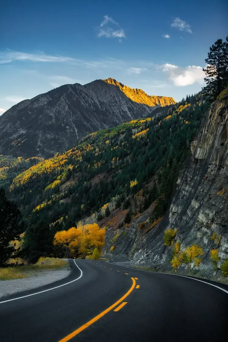 Winding mountain road with fall foliage on either side, leading through a forested valley with tall mountains in the background, under a blue sky with scattered clouds.	
