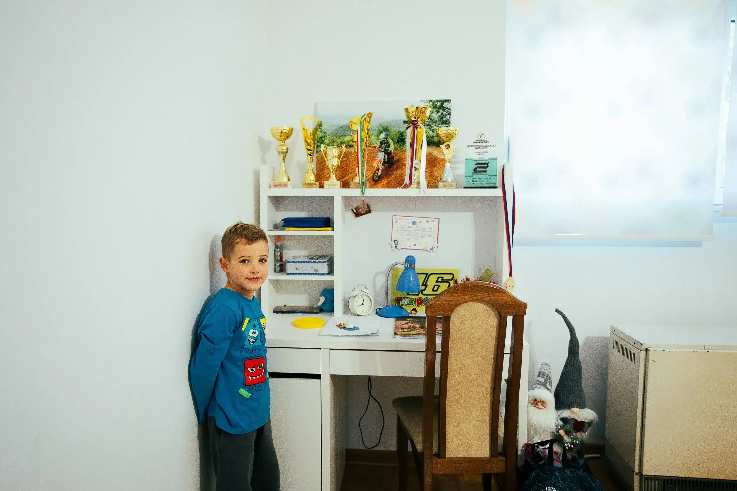 A young boy in a blue shirt standing in a cluttered room next to a desk and a wooden chair, with a shelf displaying trophies and medals behind him, and Christmas gnomes on the floor to the right.
