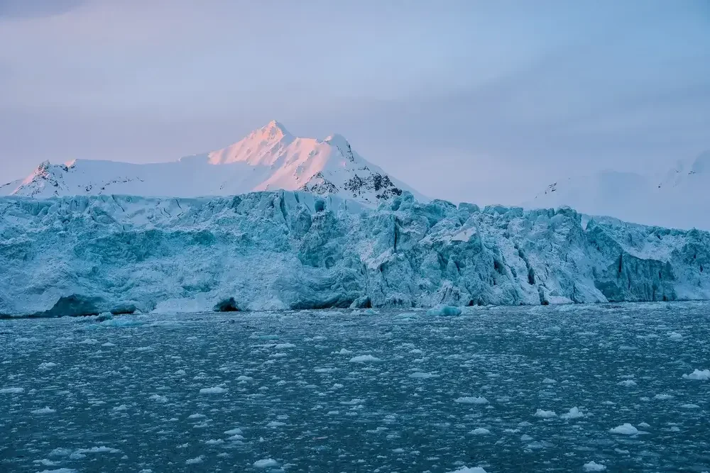A large glacier with a snow-covered mountain in the background under a pale blue sky.
