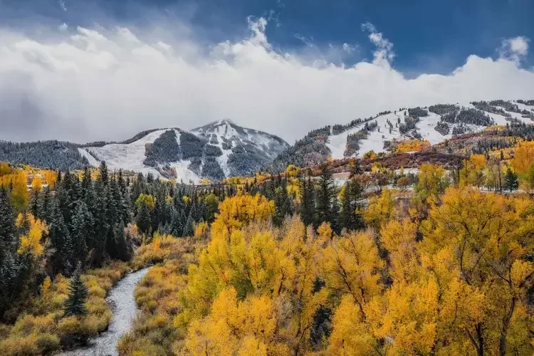 A scenic mountain landscape with snow-covered slopes, evergreen trees, and vibrant yellow and orange autumn foliage, under a partly cloudy sky.