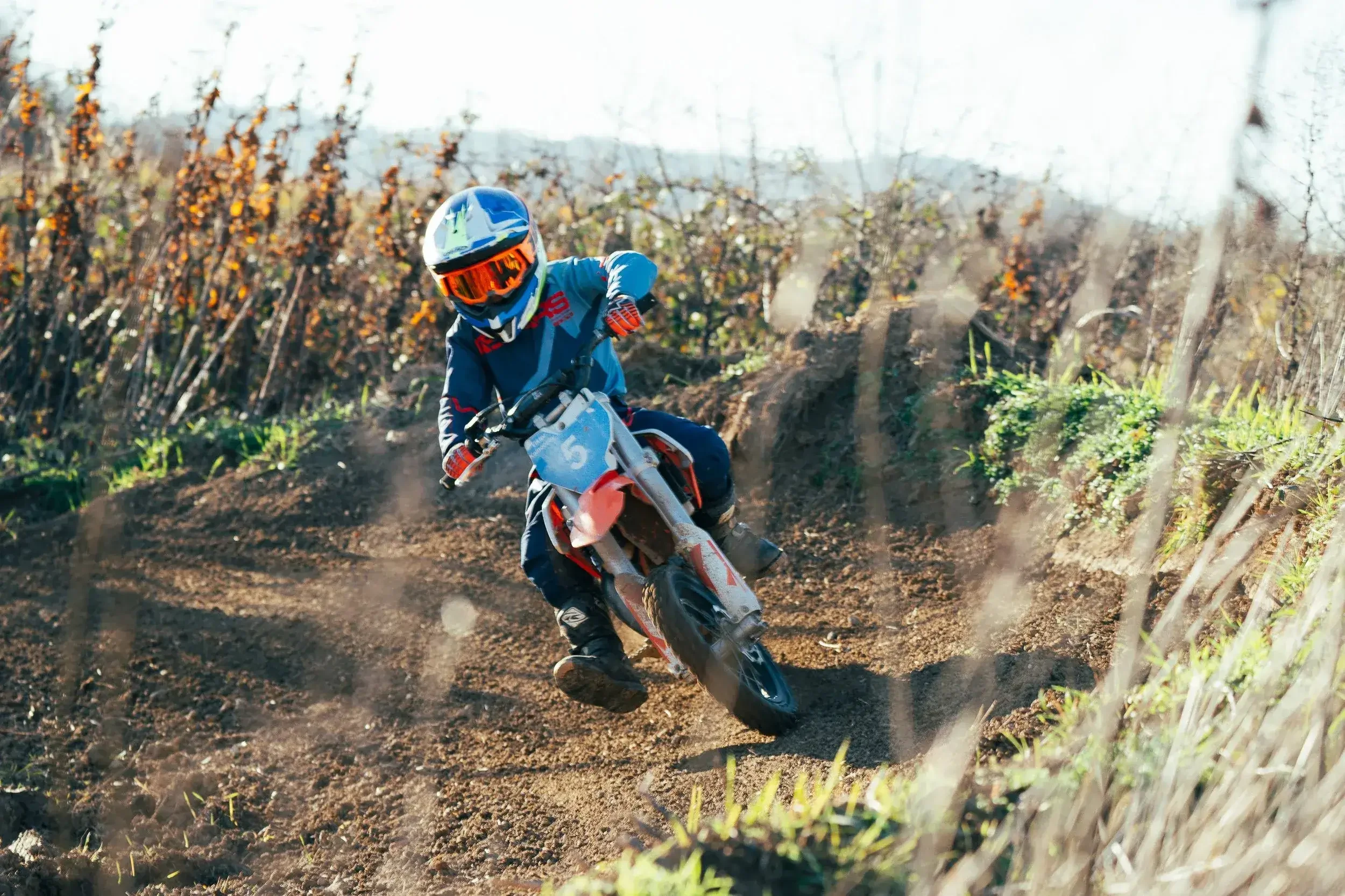 Child riding a motocross dirt bike on a dirt trail surrounded by fields and bushes.
