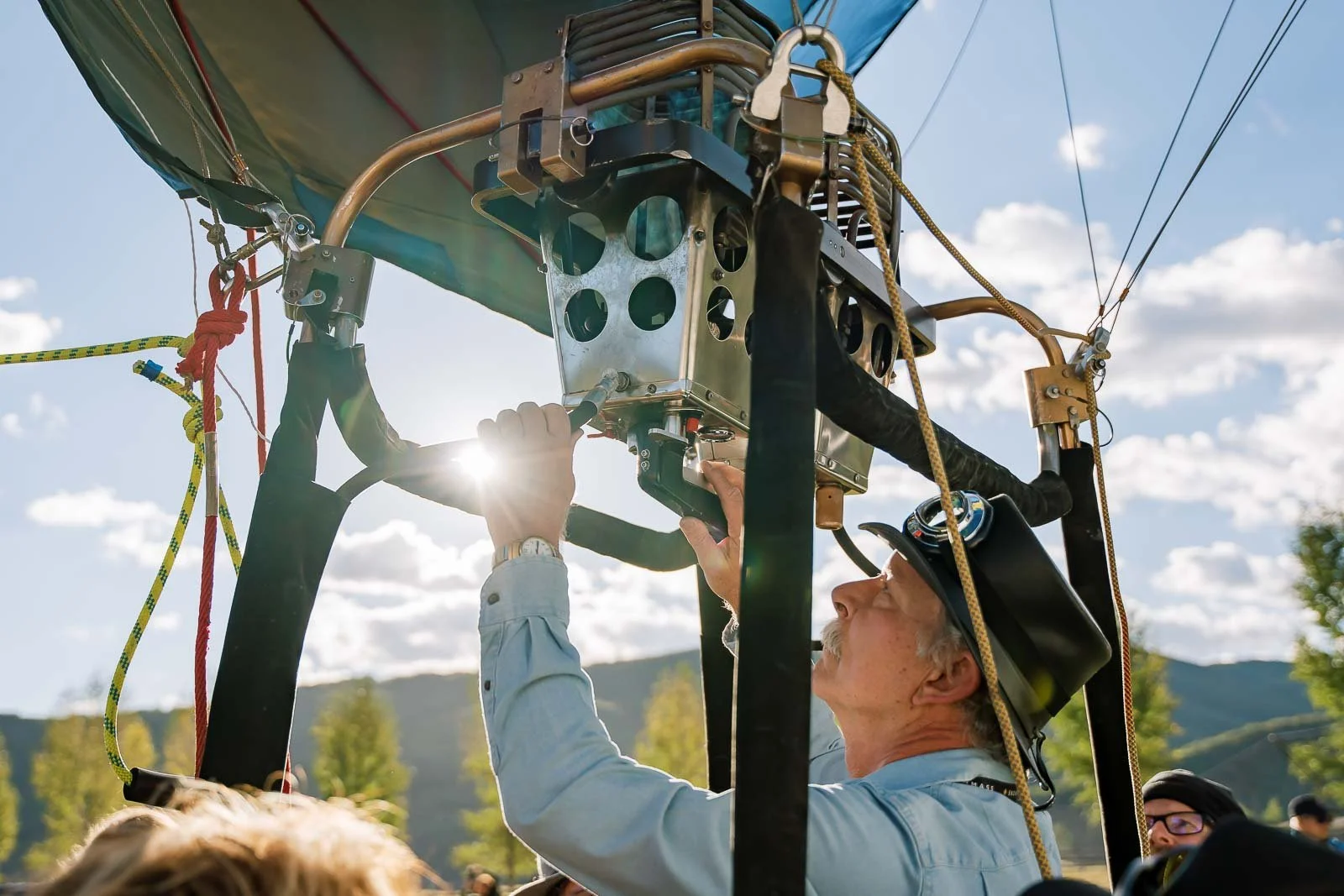 A man in a blue shirt and black hat with a headlamp is working on a hot air balloon basket's metal frame, adjusting equipment outdoors with trees and mountains in the background.