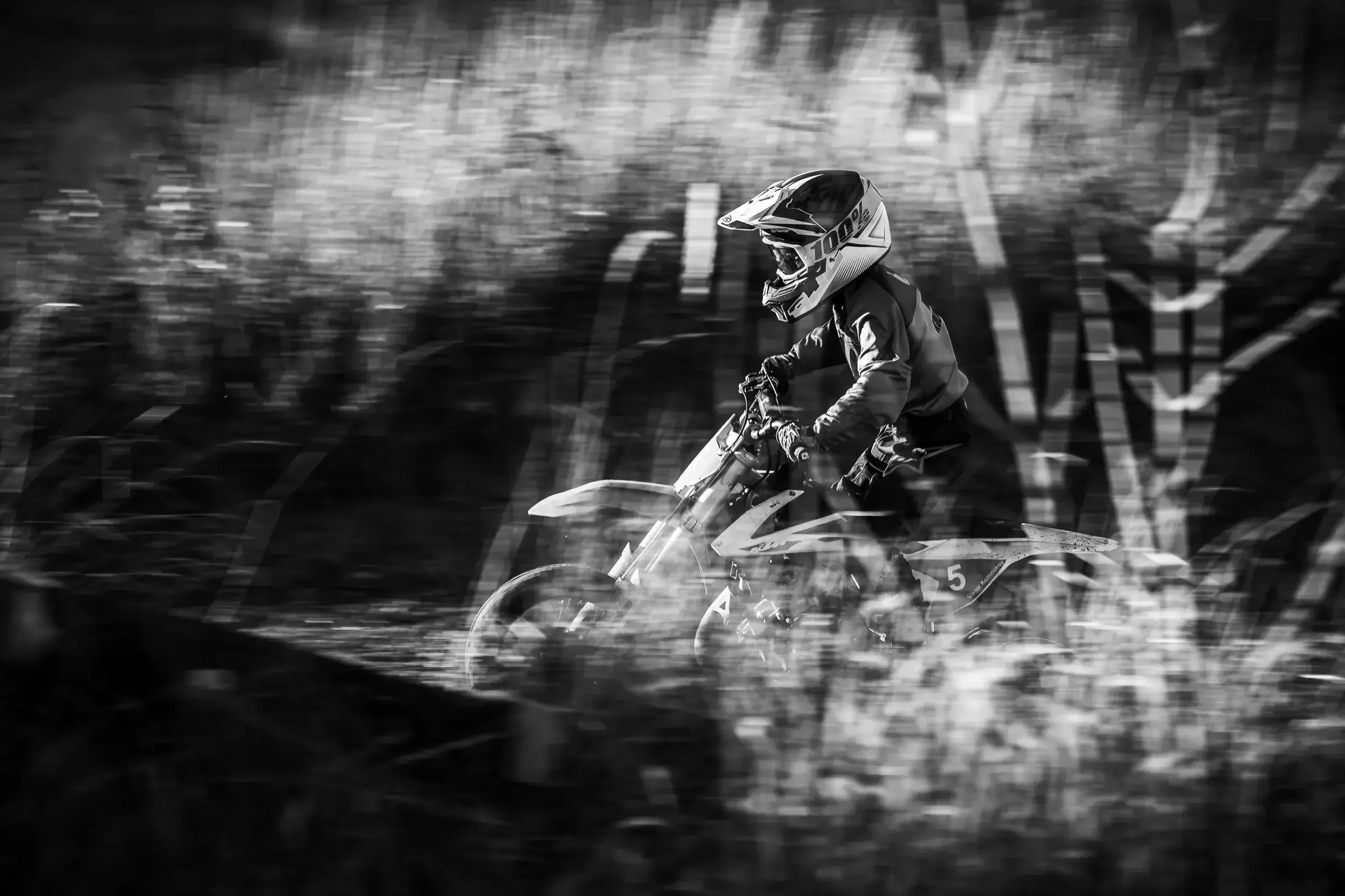 A person riding a dirt bike in motion outdoors, wearing a helmet and protective gear, with a blurred natural background in black and white.
