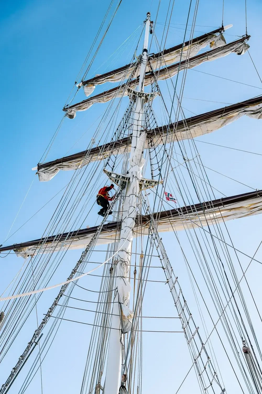 A person in red jacket climbing the mast of a tall sailing ship with multiple sails, rigging, and ropes against a clear blue sky.
