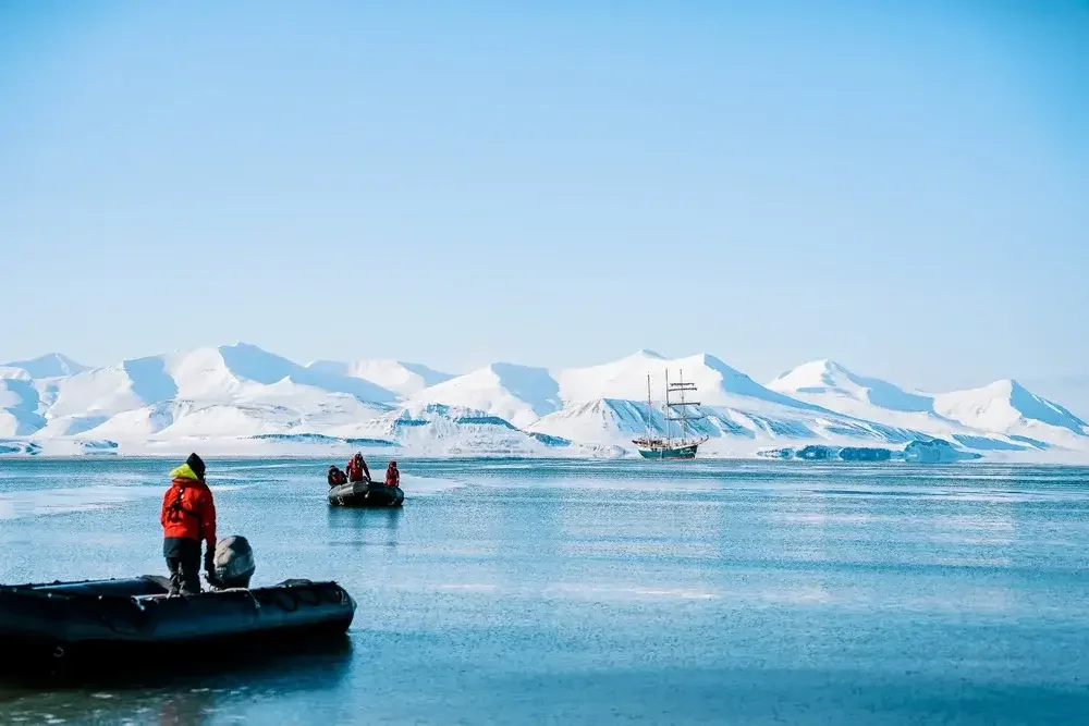 People in red jackets on inflatable boats near snowy mountains and a sailing ship in icy waters.
