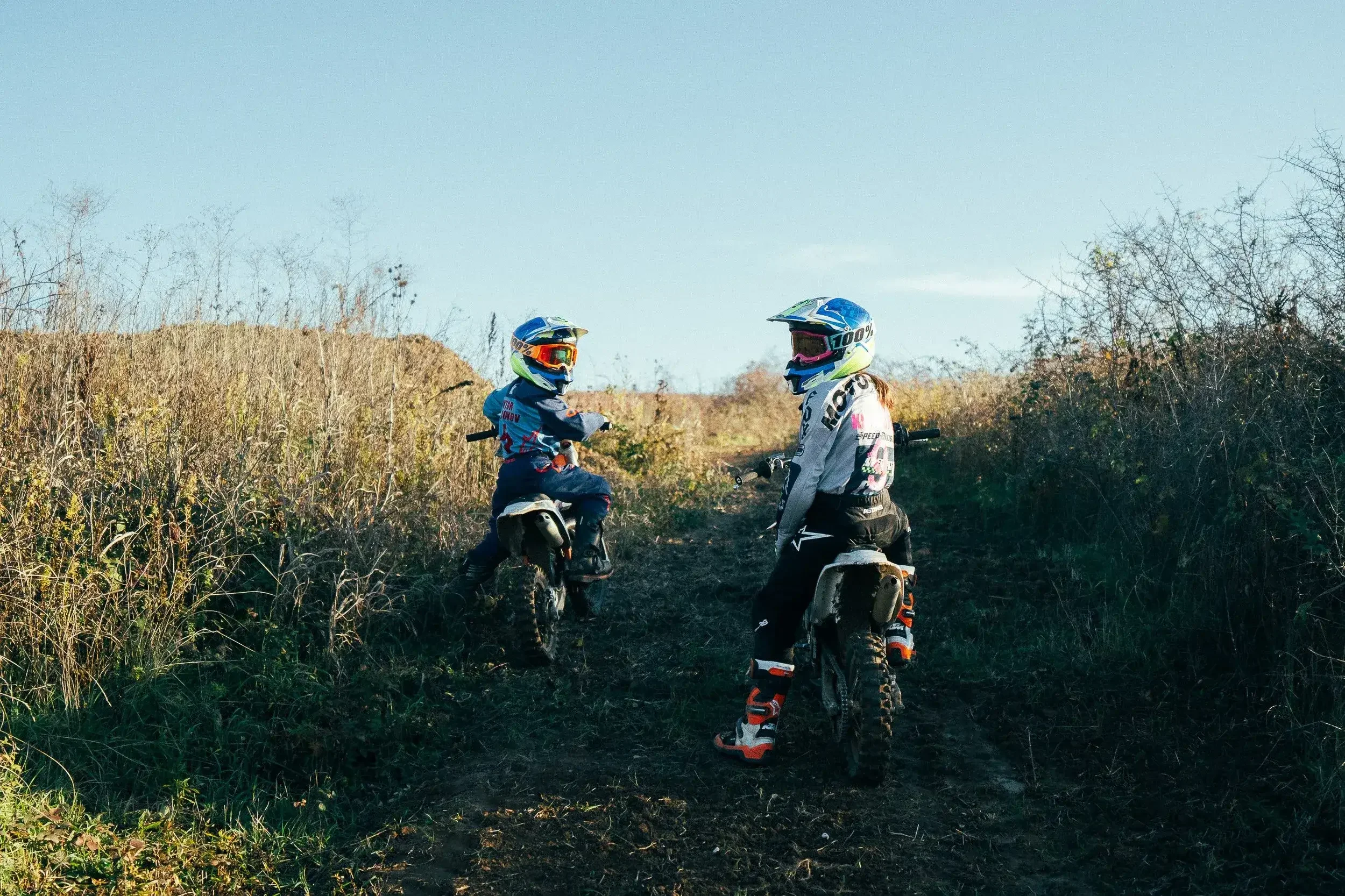 Two children wearing motocross gear and helmets, sitting on dirt bikes on a trail surrounded by dry grass and bushes during daytime.
