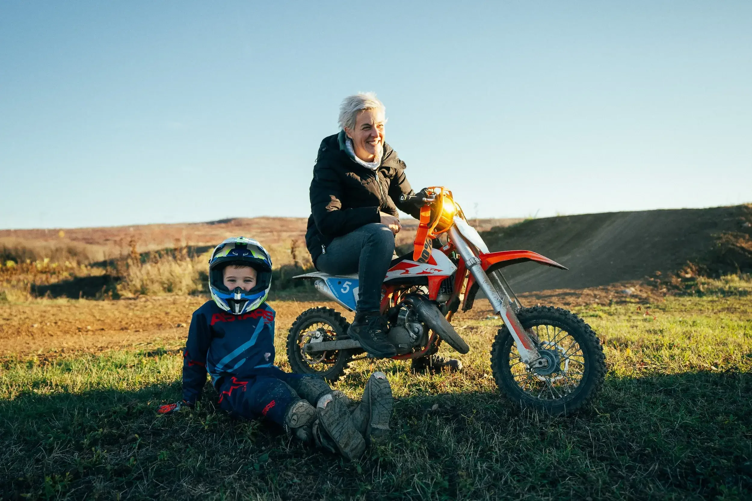 A woman riding a red dirt bike with a young boy sitting on the ground next to her in an outdoor rural setting, both wearing helmets and protective gear, during daytime with clear sky.
