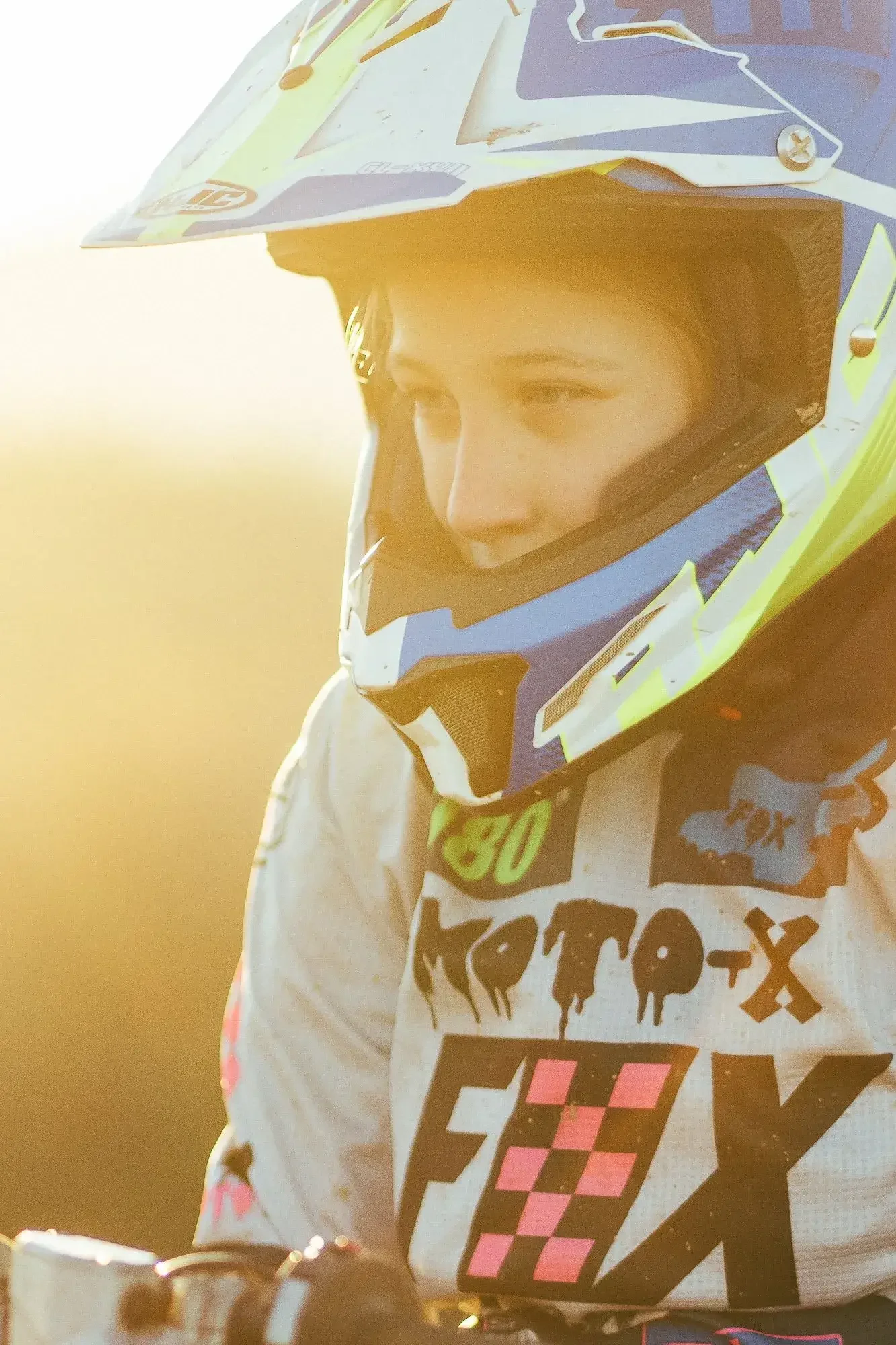 A young person wearing a motocross helmet and gear, looking focused, with sunlight shining in the background.
