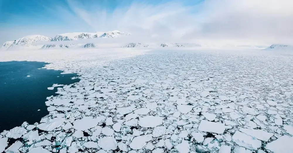 A sailboat stranded on ice-covered water in a cold, snowy landscape with mountains on both sides and a pastel sky.
