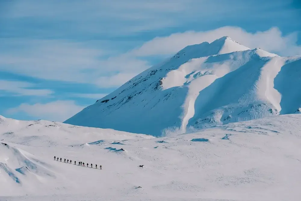 A group of hikers trekking across a snowy mountain landscape with a large snow-covered peak and blue sky in the background.
