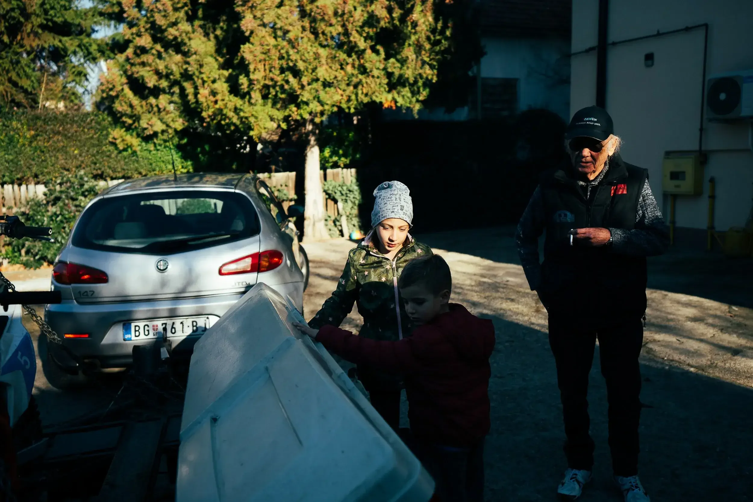 Two children and an elderly man standing outdoors near a silver car and a trailer, with trees and a building in the background, during daytime.
