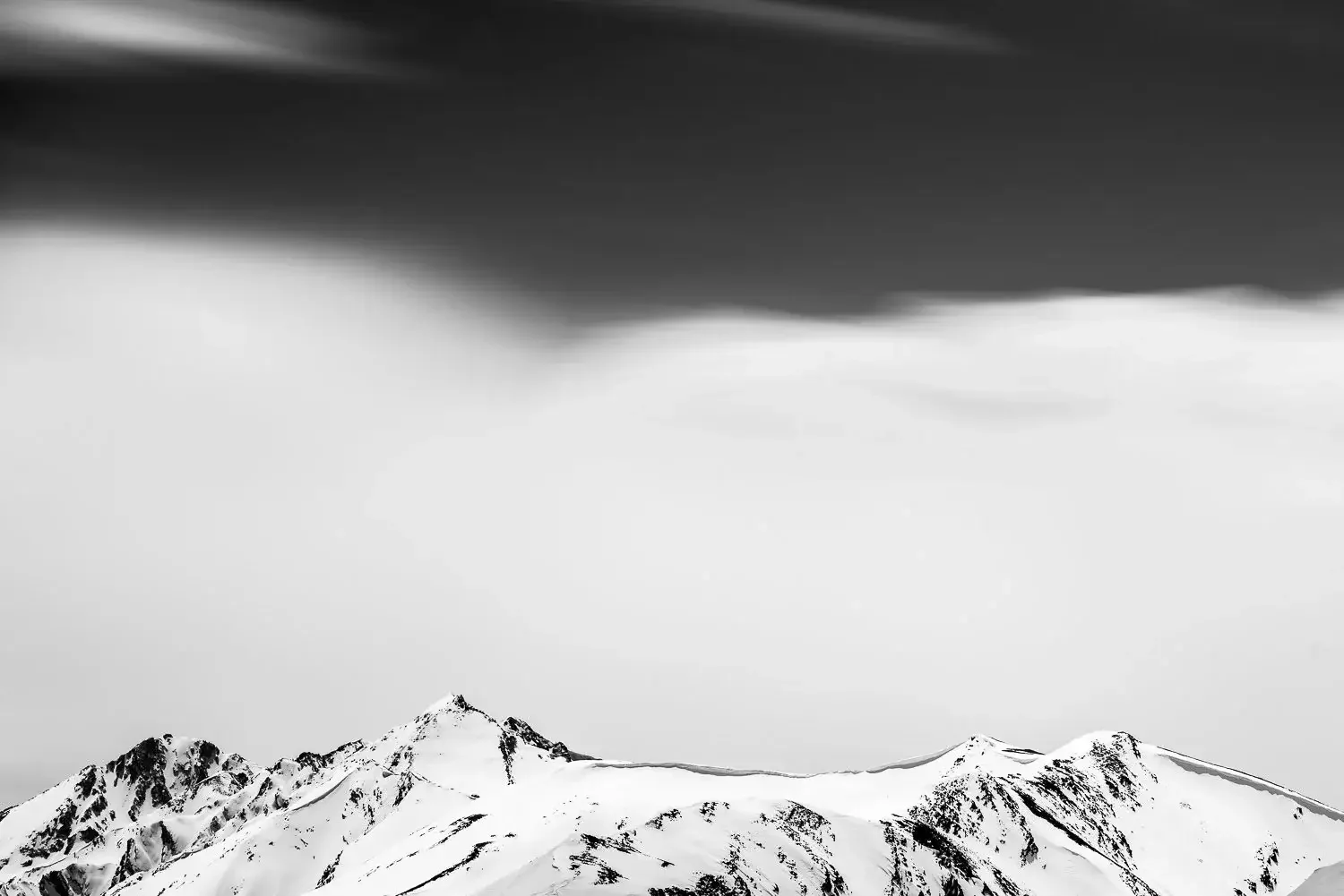 Black and white photo of snow-covered mountain peaks with dark dramatic clouds overhead.
