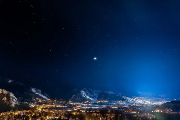 Nighttime mountain landscape with snow-covered peaks, illuminated town lights in the valley, and a starry sky with a bright planet or star.
