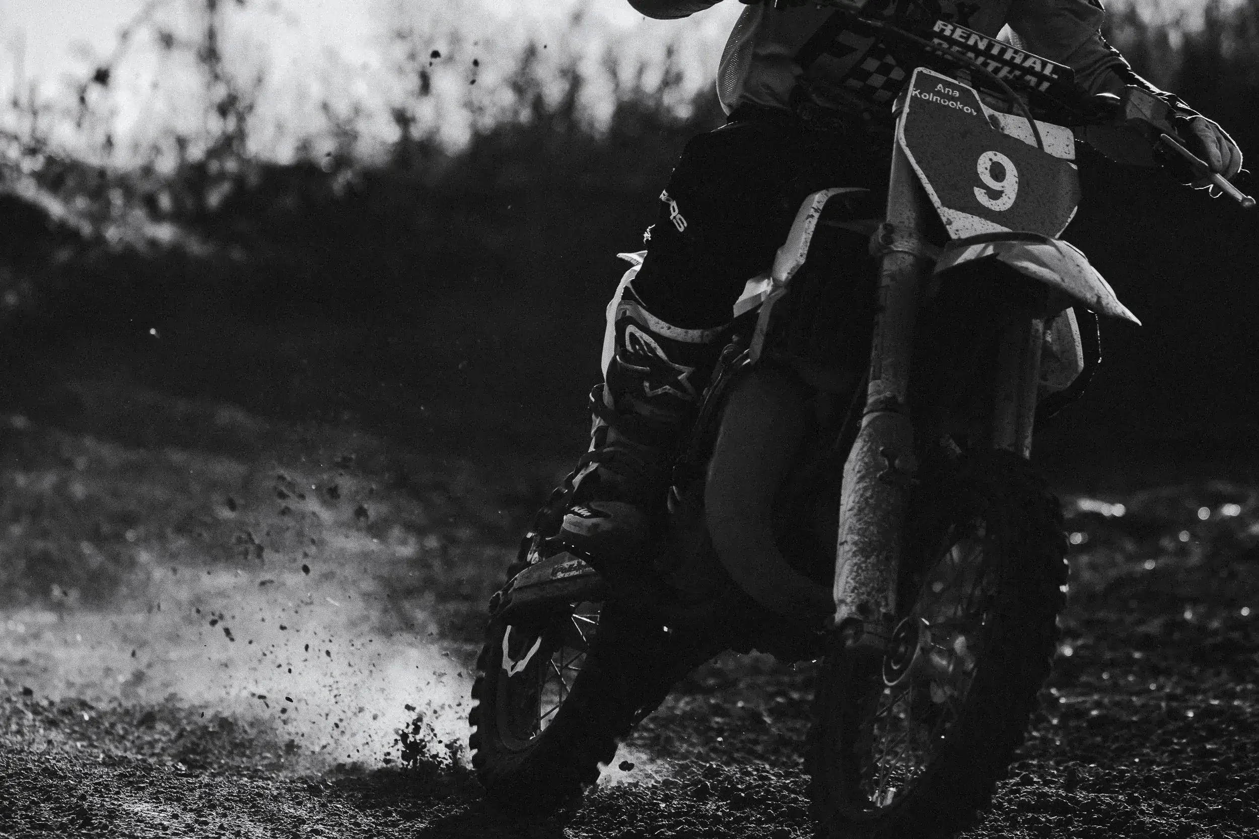 A motocross rider on a dirt bike speeding through a muddy track, kicking up dirt and debris in black and white.

