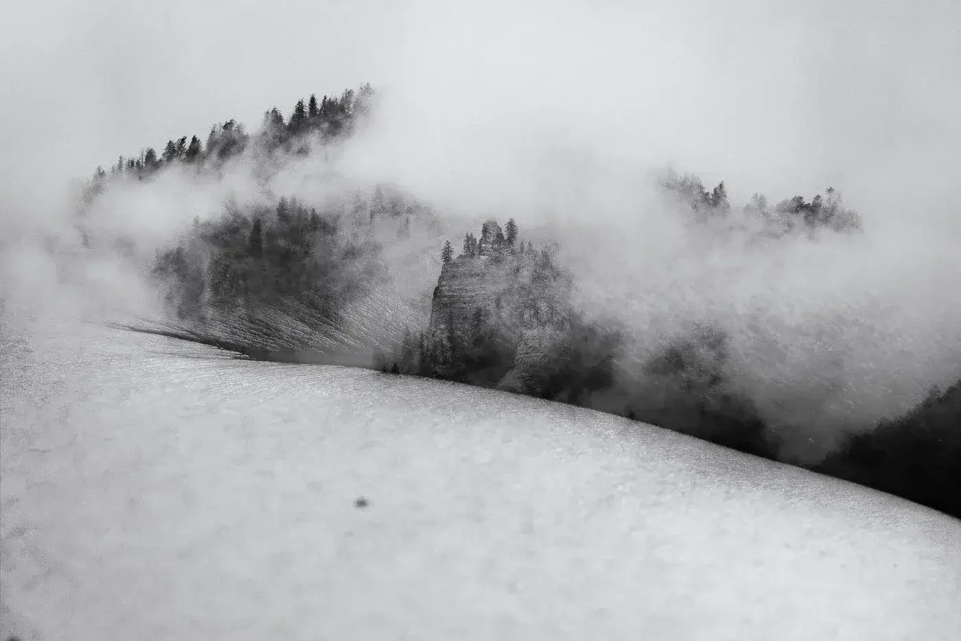 Black and white photograph depicting a mountain landscape with fog and trees, creating a mysterious and atmospheric scene.
