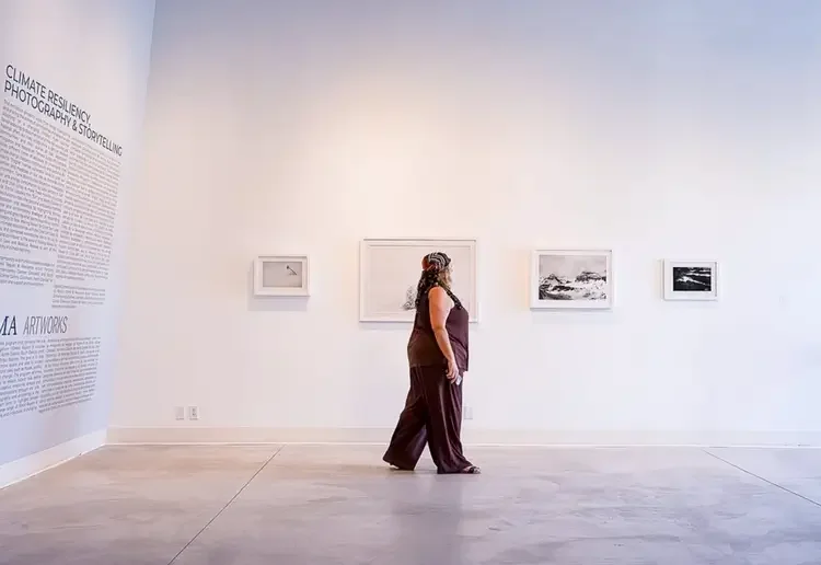 A woman in a maroon outfit and colorful headscarf standing in a white art gallery looking at black and white landscape photographs on the wall.