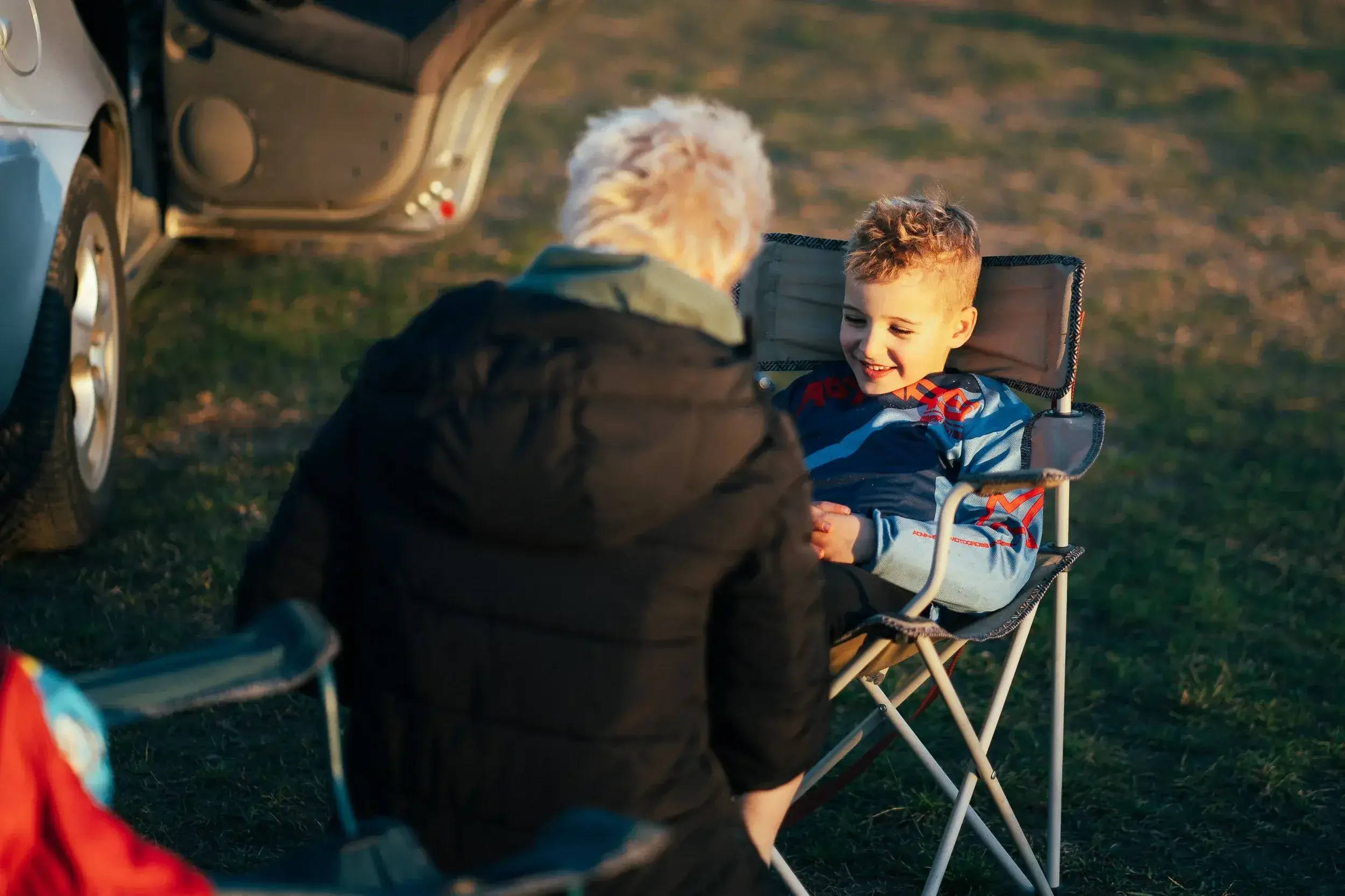 A young boy sitting in a portable camping chair outdoors, smiling and talking with an elderly woman with white hair, near a parked vehicle on grassy terrain during sunset.
