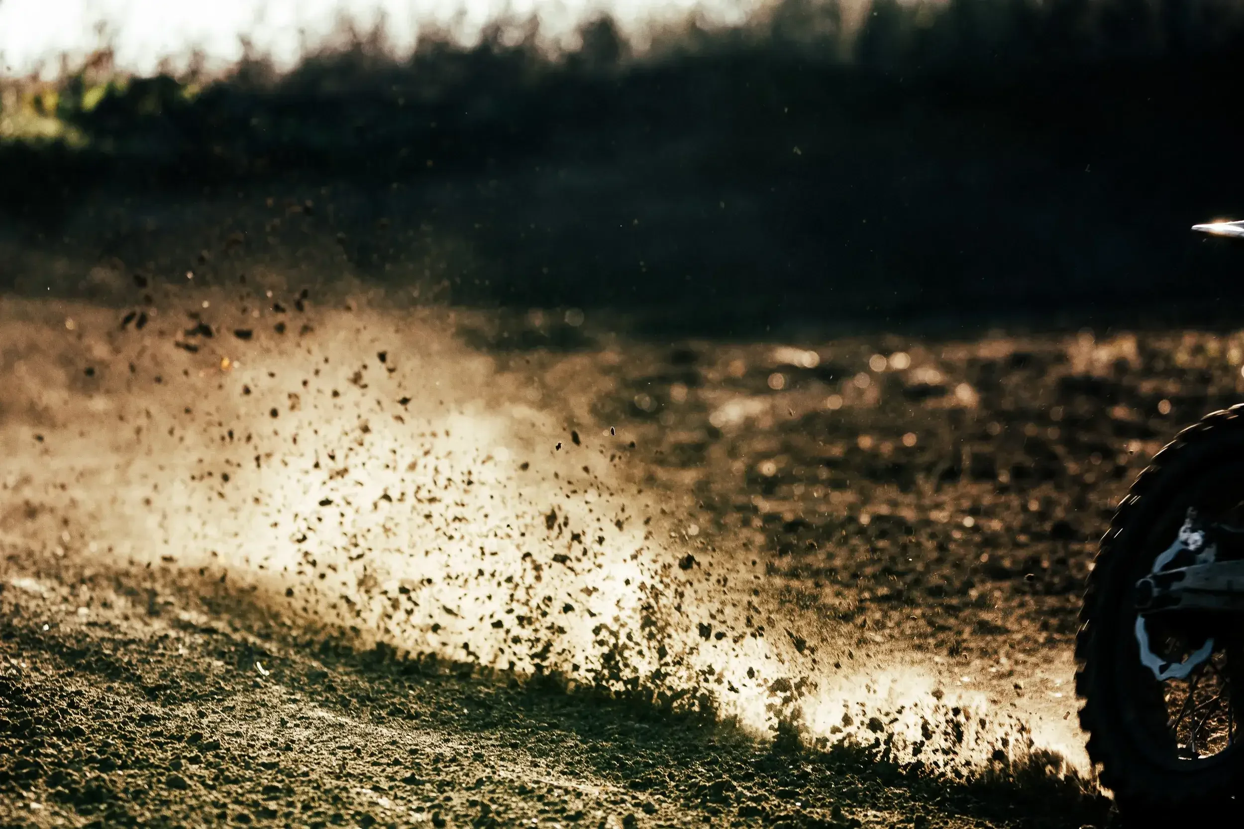 Off-road dirt bike kicking up dirt dust on a trail with a motorcycle wheel visible on the right side.
