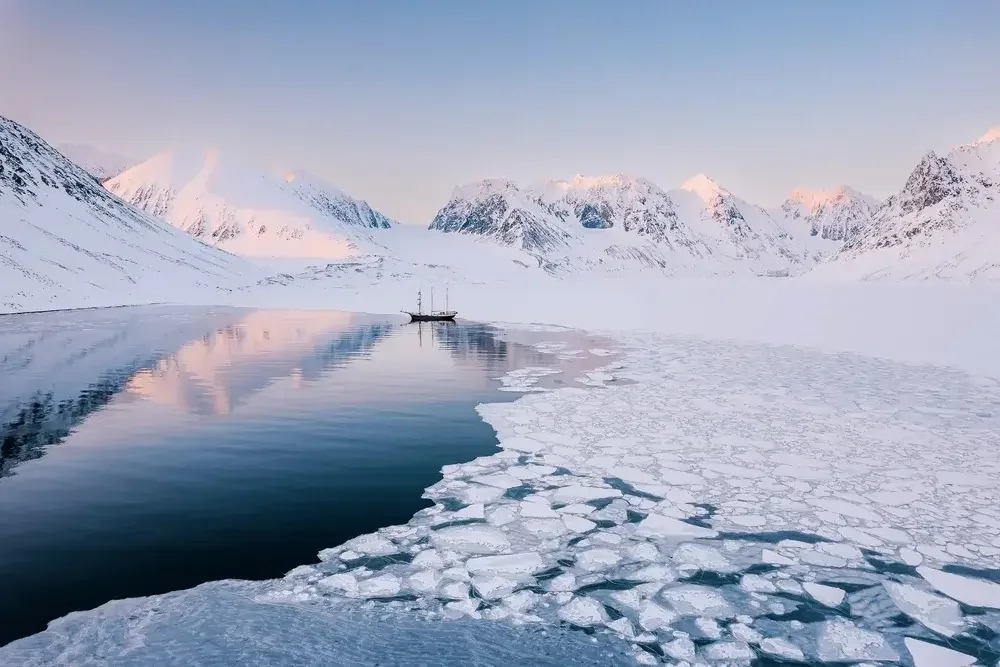 A snowy mountain landscape with a partly frozen body of water and a sailboat in the distance.