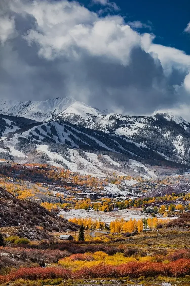 Spectacular mountain landscape with snow-capped peaks, colorful fall foliage, and a cloudy sky.
