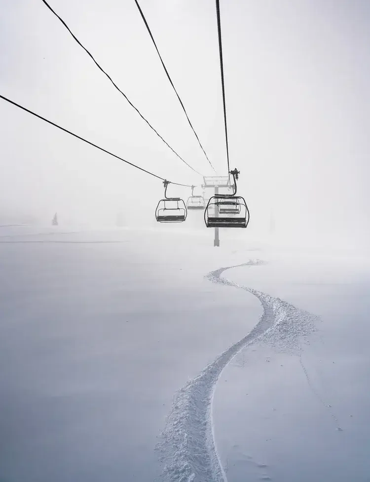 Empty ski lift chairs suspended over a snow-covered landscape with ski tracks and foggy background.
