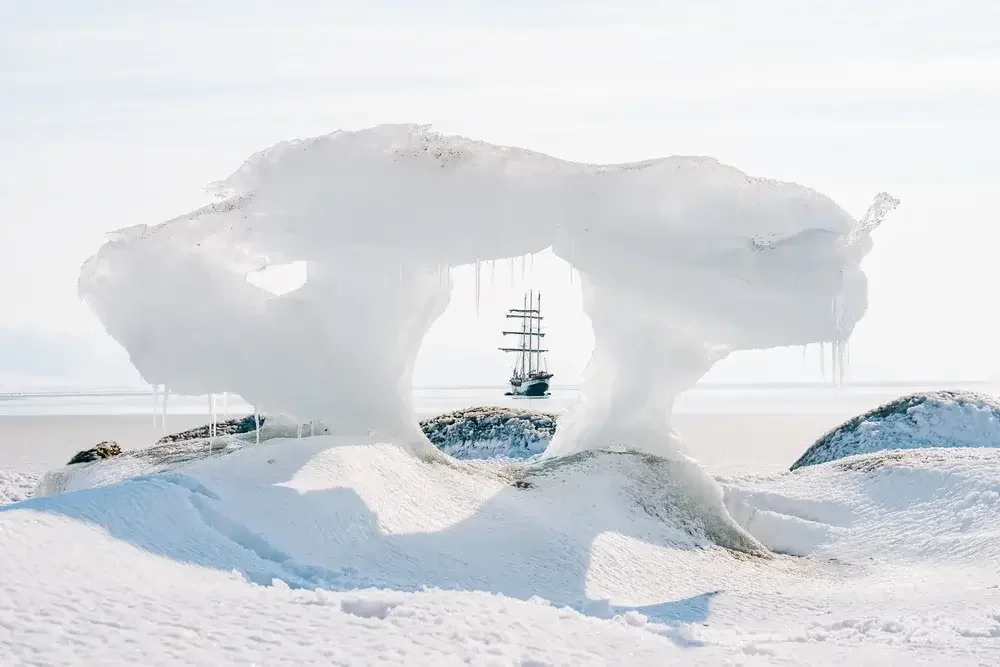 A ship seen through a hole in a large, icy snow formation on a snowy landscape with a body of water in the background.
