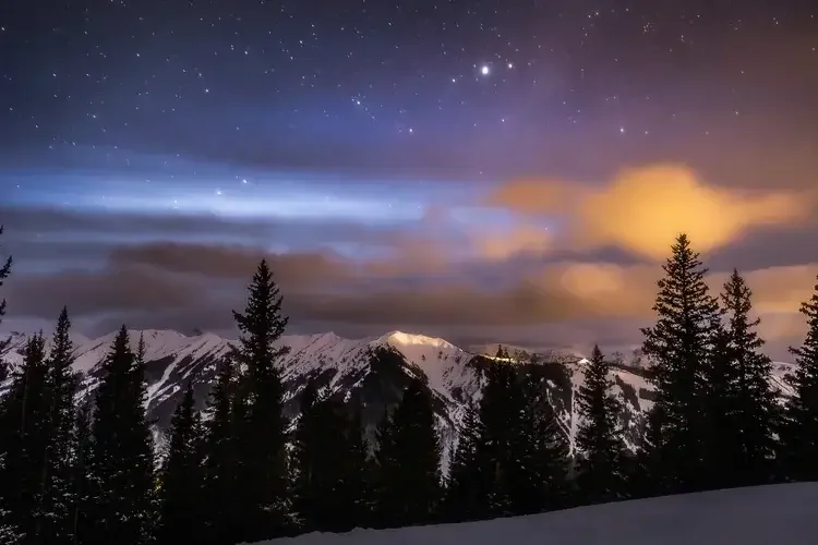 Night sky filled with stars and the Milky Way visible over a forested area with a rocky river in the foreground.
